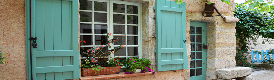 A photo of a house in France with flowers in the window ledges. The doors and windows of the property are painted in a teal blue.